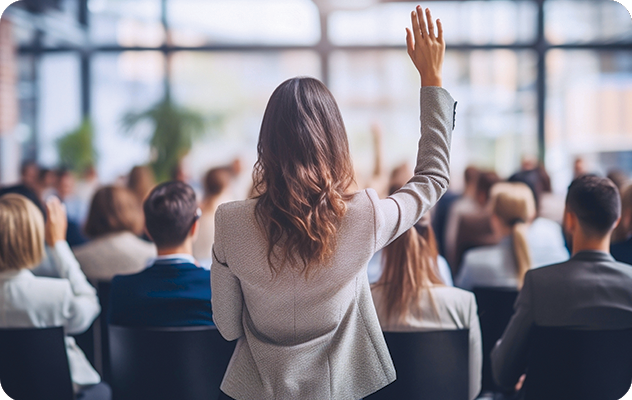 A picture of the back of a woman standing a raising her hand in an audience at a conference.