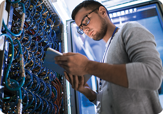 A picture of a young IT professional in glasses accessing information on a tablet in front of a bank of servers.