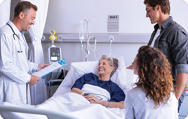 A picture of a young doctor talking to an older female patient smiling in her bed with a family member and a qualified medical interpreter present