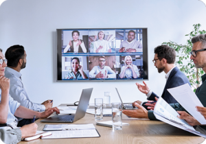 A picture of a large corporate conference room holding a meeting with people in person and on the screen virtually.