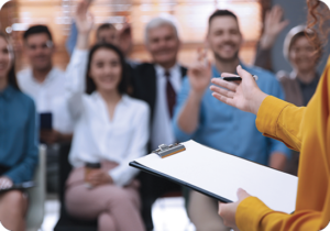 A picture of adult age students raising their hands for a teacher with a clipboard.