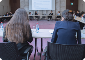 A picture of two people sitting in the foreground of a large square meeting space framed by tables during a government meeting.