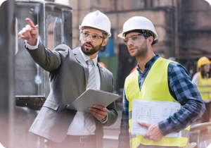 A picture of a man in a suit and a hardhat pointing out something in a factory to another man in a safety vest