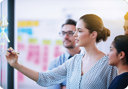 A picture of a woman organizing writing on sticky notes on a wall explaining process to two other employees during the interpreting and translation process.